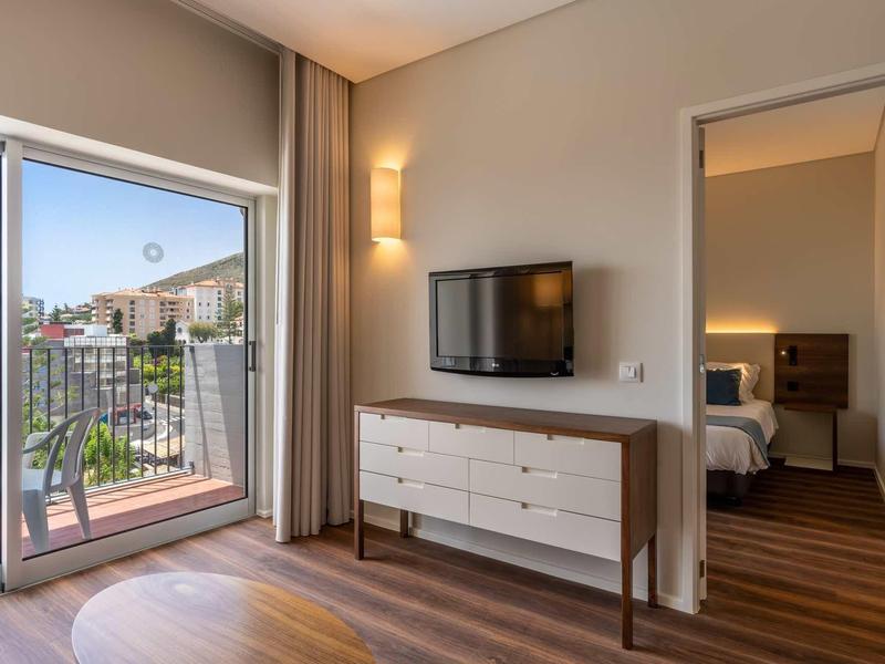 Helles Wohnzimmer mit Holzboden, TV auf Kommode und Balkon mit Blick auf Nachbarhäuser.