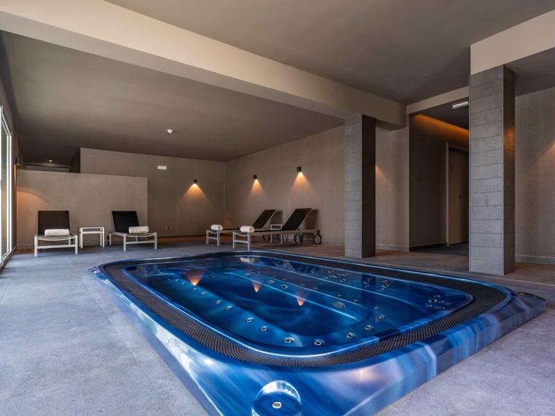 Moderner Innenpool mit blauem Whirlpool, grauen Wänden und gemütlichen Liegestühlen am Fenster.