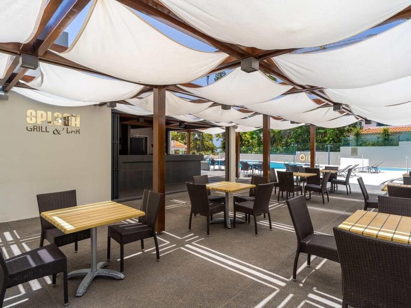 Überdachter Außenbereich mit Tischen und Stühlen aus dunklem Material, heller Boden und Sonnensegel.