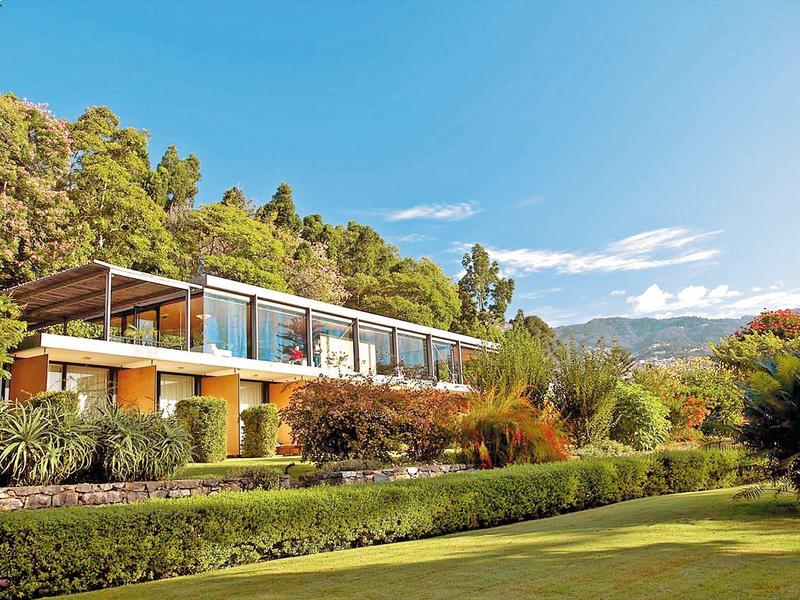 Hotel moderno con grande facciata in vetro circondato da verde rigoglioso e montagne sullo sfondo.