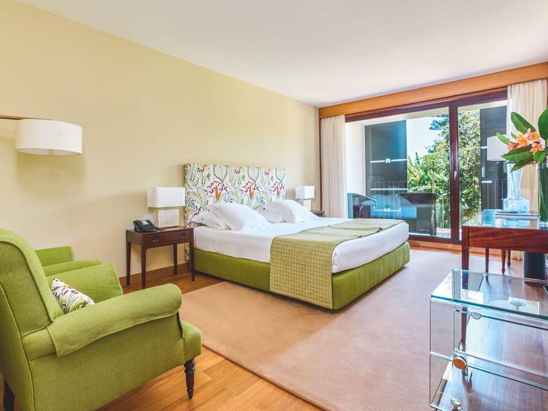 Camera d'hotel luminosa con letto matrimoniale, poltrona verde e accesso al balcone con piante.