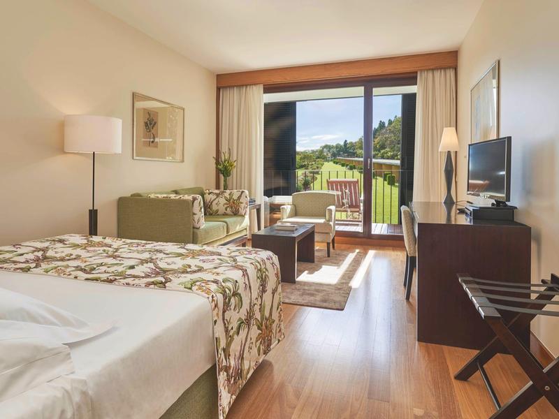 Camera d'hotel luminosa con letto matrimoniale, divano e balcone con vista sulle montagne.