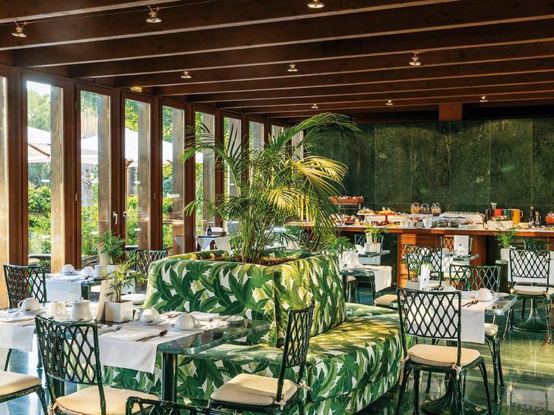 Area ristorante dell'hotel moderna con grandi finestre, piante e posti a sedere eleganti.