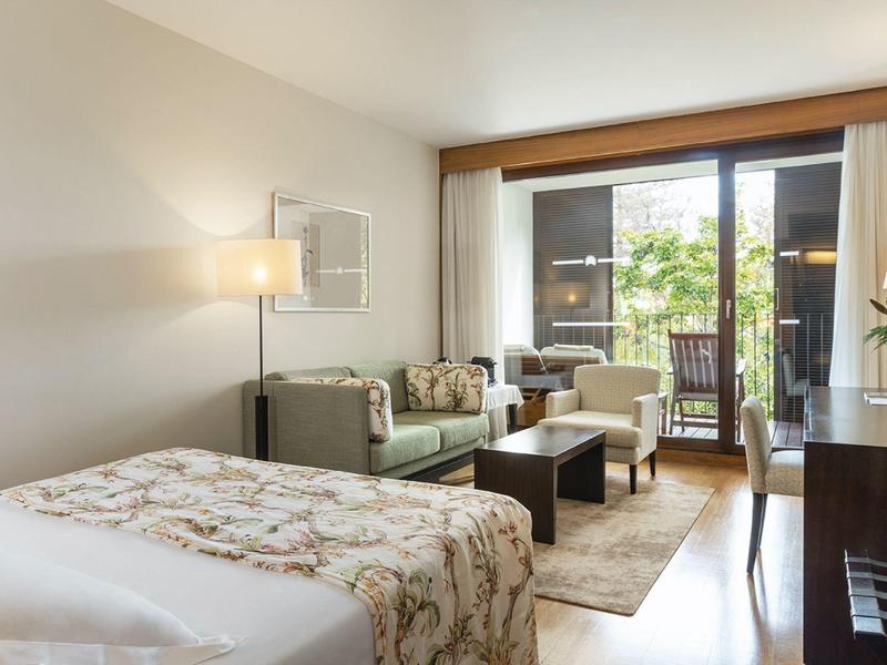 Camera d'hotel luminosa con letto, divano, tavolino e balcone con vista su area verde.