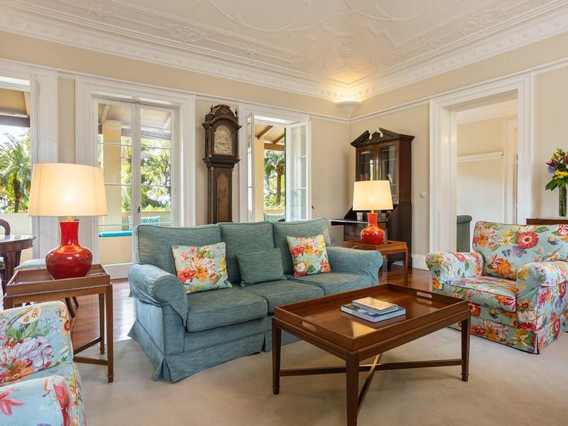 Soggiorno luminoso con divano blu, poltrona floreale e mobili in legno accanto a grandi finestre.