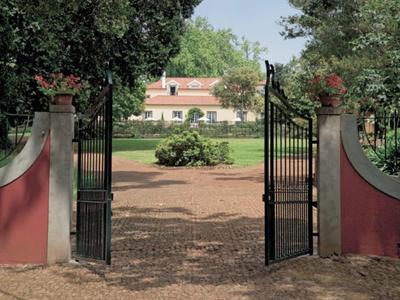 Otwarta żelazna brama prowadzi do zadbanego majątku z białym domem i ogrodem.