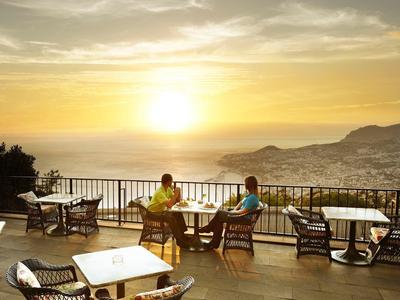 Dwie osoby siedzą na tarasie z krzesłami i stołami, z widokiem na morze o zachodzie słońca.