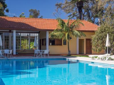 Widok na spokojny basen z leżakami przed żółtym domem i drzewami w tle.