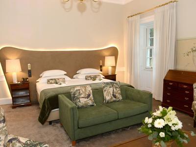 Przytulny pokój hotelowy z łóżkiem podwójnym, zieloną kanapą i kwiatową dekoracją w ciepłym świetle.