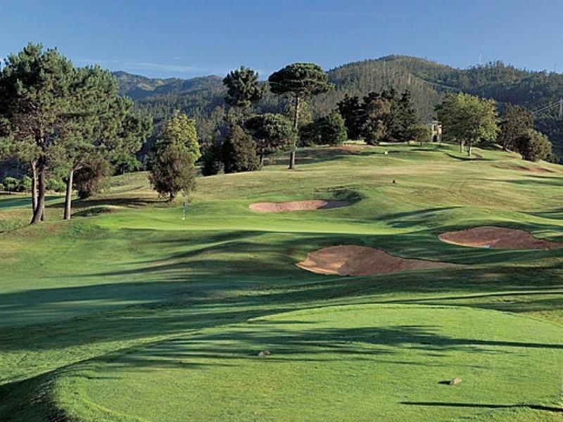 Zielone pole golfowe z oczkiem wodnym, drzewami i widokiem na morze w tle.