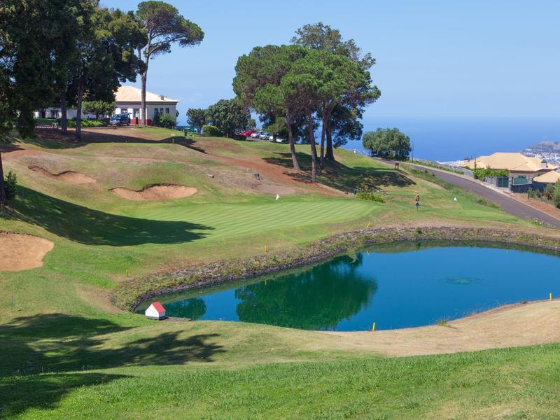 Zielone pole golfowe z oczkiem wodnym, drzewami i widokiem na morze w tle.