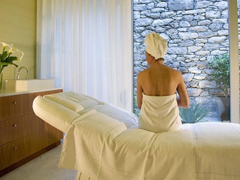 Pokój spa z leżanką, na której siedzi kobieta owinięta w ręcznik, patrząca przez szklane okno na ogród.