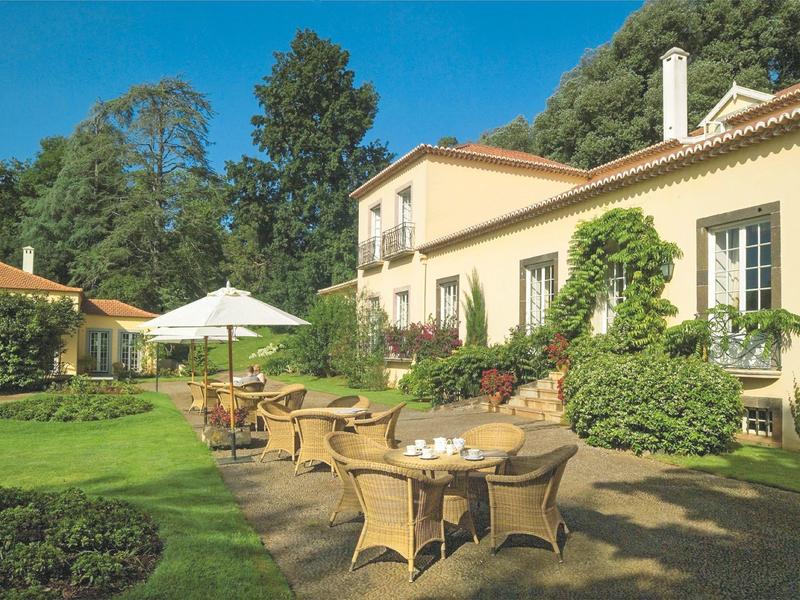 Ogród z meblami i parasolem przed żółtym domem na zielonym tle.