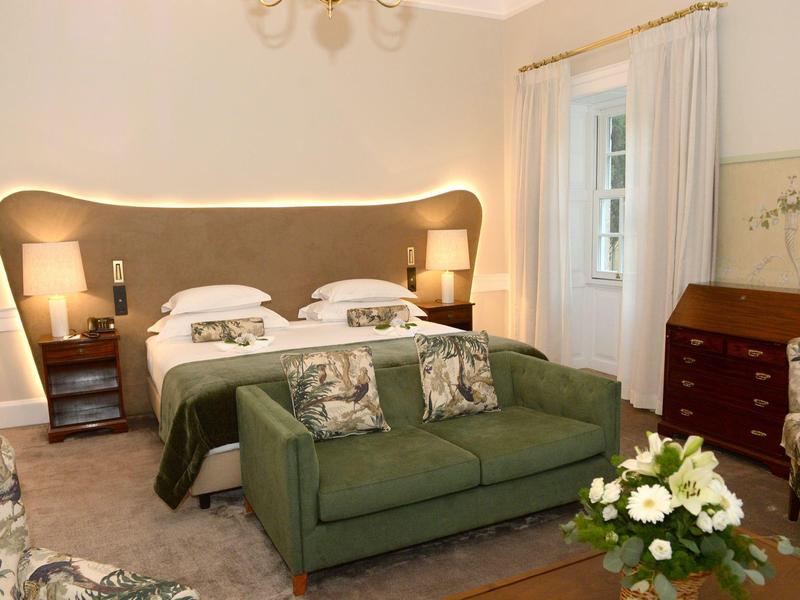 Przytulny pokój hotelowy z łóżkiem podwójnym, zieloną kanapą i kwiatową dekoracją w ciepłym świetle.