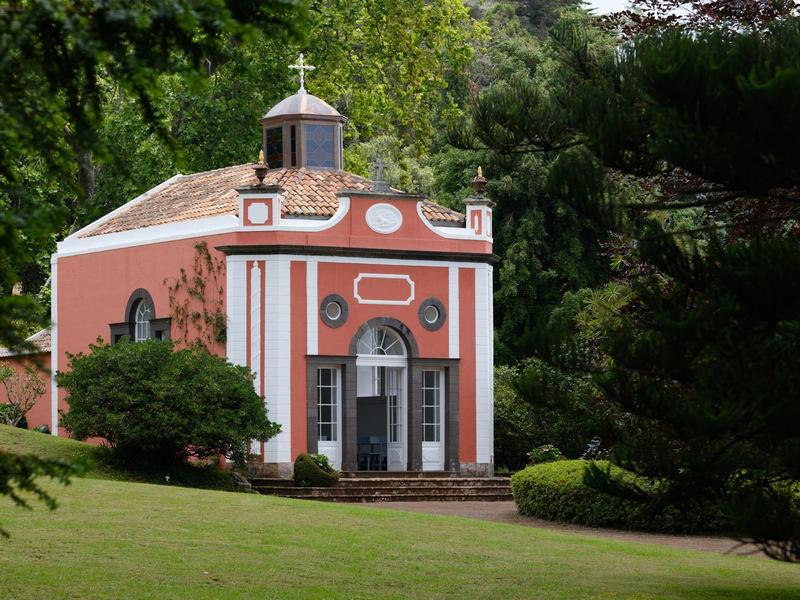 Mały czerwony budynek z białymi kolumnami i krzyżem na dachu w zielonym parku