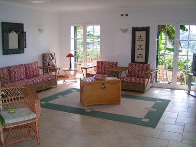 Helles Wohnzimmer mit geflochtenen Möbeln, roten Polstern, Holzkiste und großen Fensterfronten.