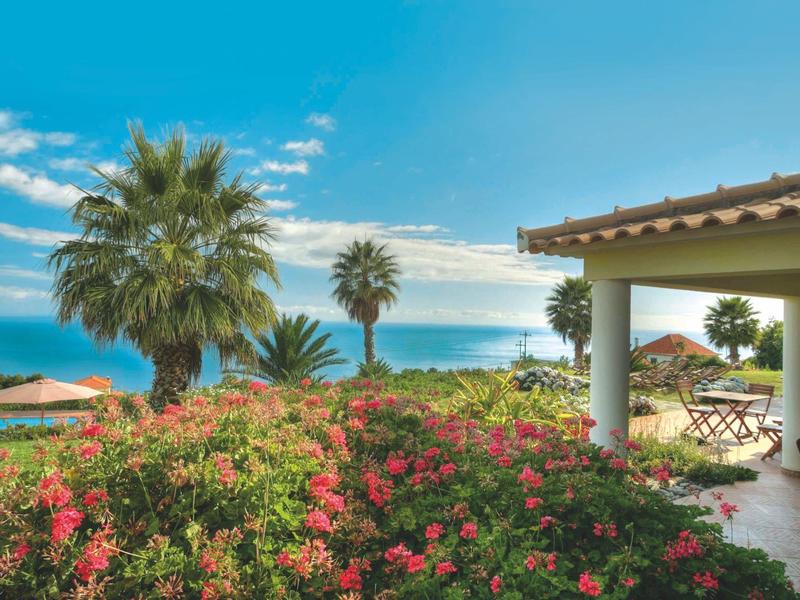 Terrace with seating area next to blooming garden and sea view under blue sky.
