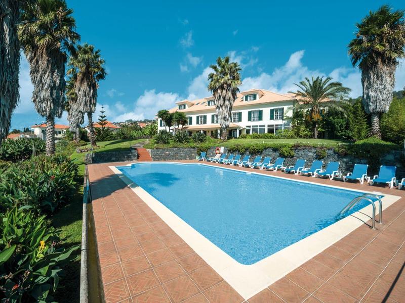 Large outdoor pool with blue lounge chairs in front of a hotel building and palm trees.
