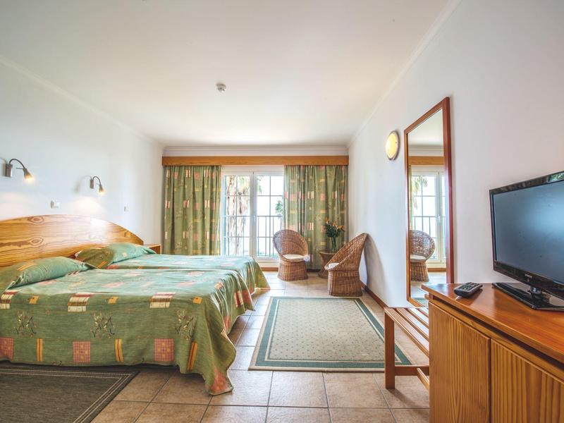 Bright hotel room with double bed, two armchairs, small rug, and TV on wooden cabinet.
