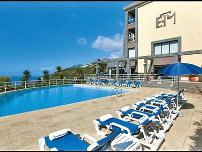 Ein blauer Pool mit weißen Liegestühlen vor einem mehrstöckigen Gebäude und klarer Himmel.