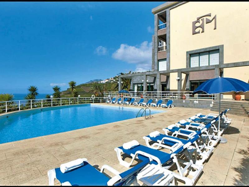 Ein blauer Pool mit weißen Liegestühlen vor einem mehrstöckigen Gebäude und klarer Himmel.