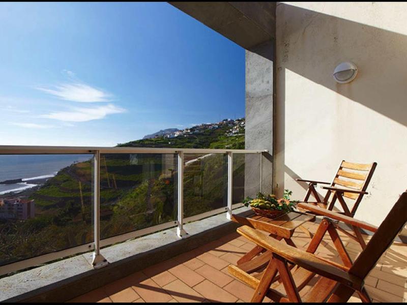 Balkon mit Holzstühlen und Tisch, Glasgeländer, Blick auf Hügel und Meer bei klarem Himmel.