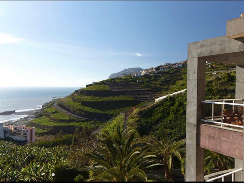 Grüne Terrassenlandschaft an Küste mit blauem Himmel und Gebäudeecke rechts im Vordergrund.
