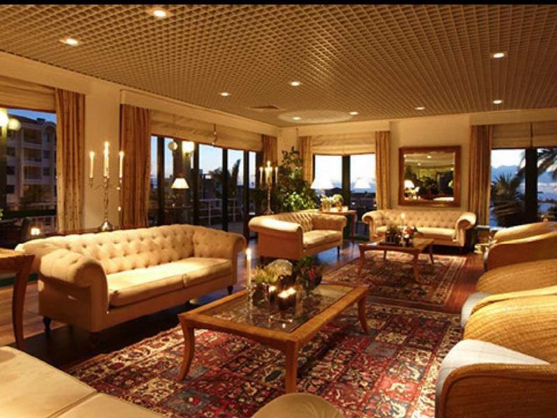 Wohnzimmer mit beigen Sesseln, Sofas, Holztischen, Teppich und warmem Deckenlicht.