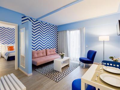 Suite d'hotel moderna con parete a zigzag blu, divano rosa e zona pranzo con vista sulla città.