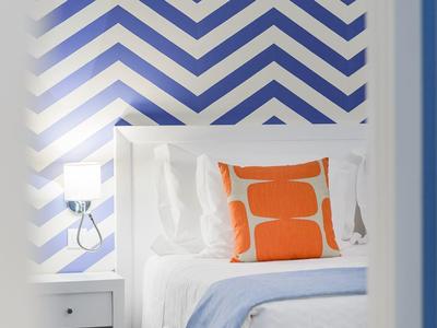 Letto con biancheria bianca, cuscino arancione, coperta blu e parete a motivo chevron.