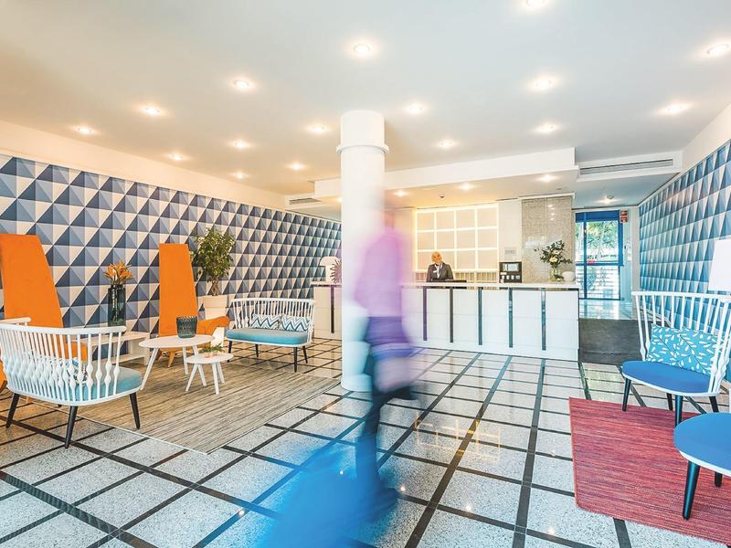 Hall dell'hotel con pareti geometriche, sedie bianche, accenti arancioni e figura sfocata in movimento.