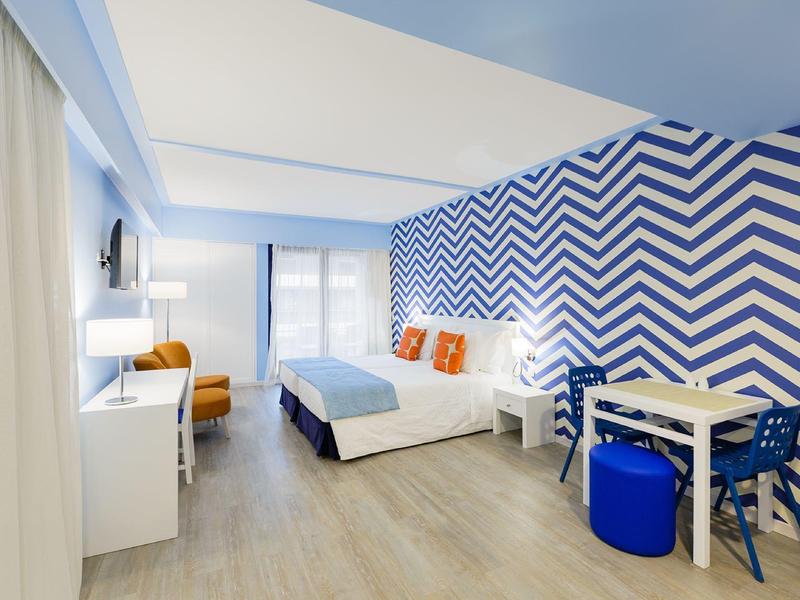 Camera d'hotel luminosa con parete a zigzag blu e bianca, letto king e zona salotto.