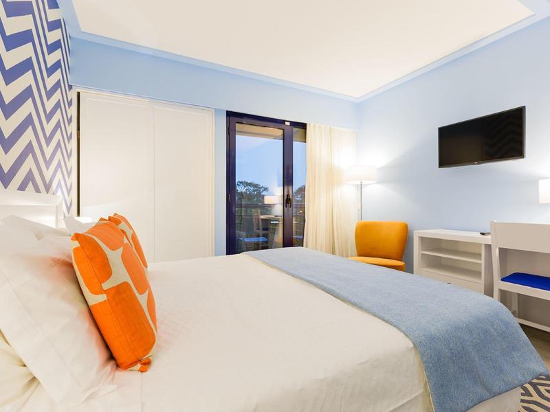 Camera d'hotel moderna con letto bianco, cuscini arancioni, coperta blu e balcone.