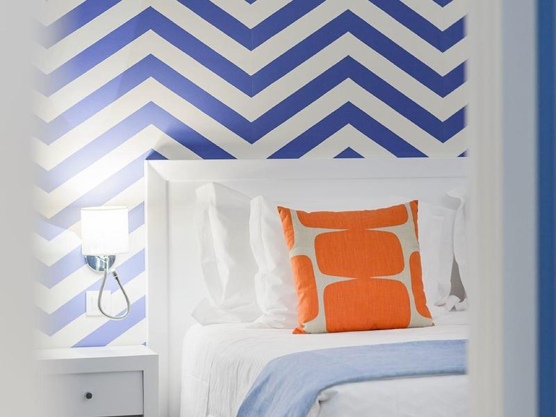 Letto con biancheria bianca, cuscino arancione, coperta blu e parete a motivo chevron.