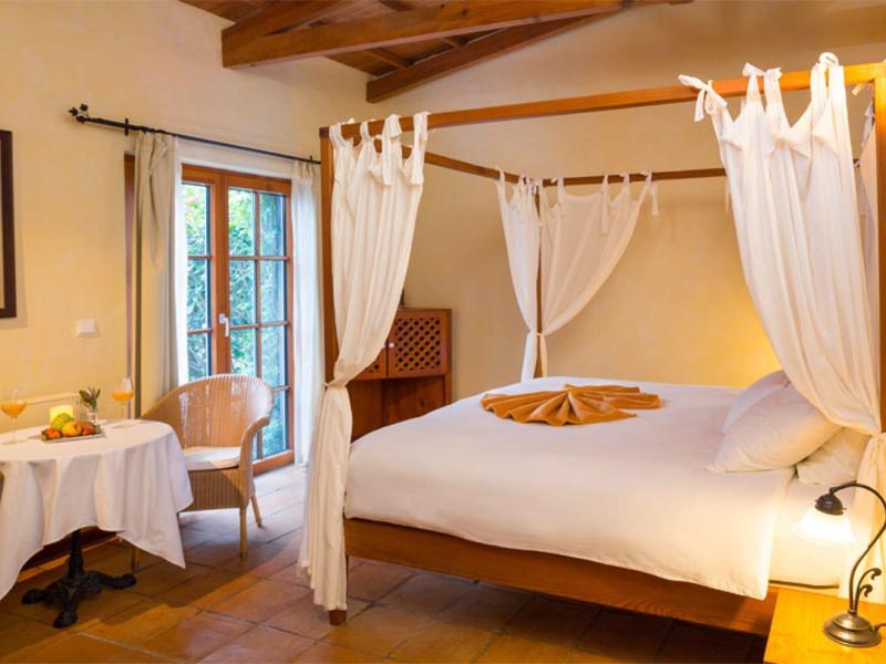 Příjemný hotelový pokoj s postelí s baldachýnem, dřevěnými trámy a malým stolkem na snídani u okna.