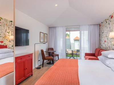 Bright hotel room with floral accent, desk, TV, and seating area with balcony access.