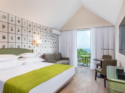 Modern hotel room with double bed, sofa, desk, and balcony overlooking the sea.