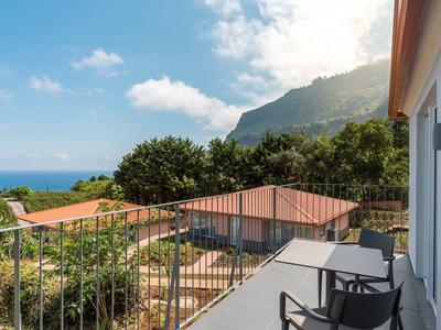 Balcony with chairs and table overlooking sea, mountains, and green vegetation on a sunny day.