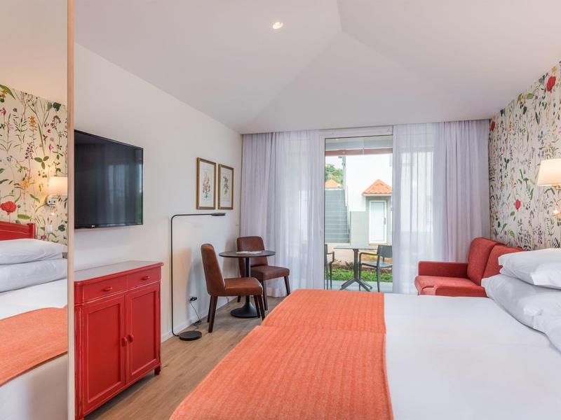 Bright hotel room with floral accent, desk, TV, and seating area with balcony access.