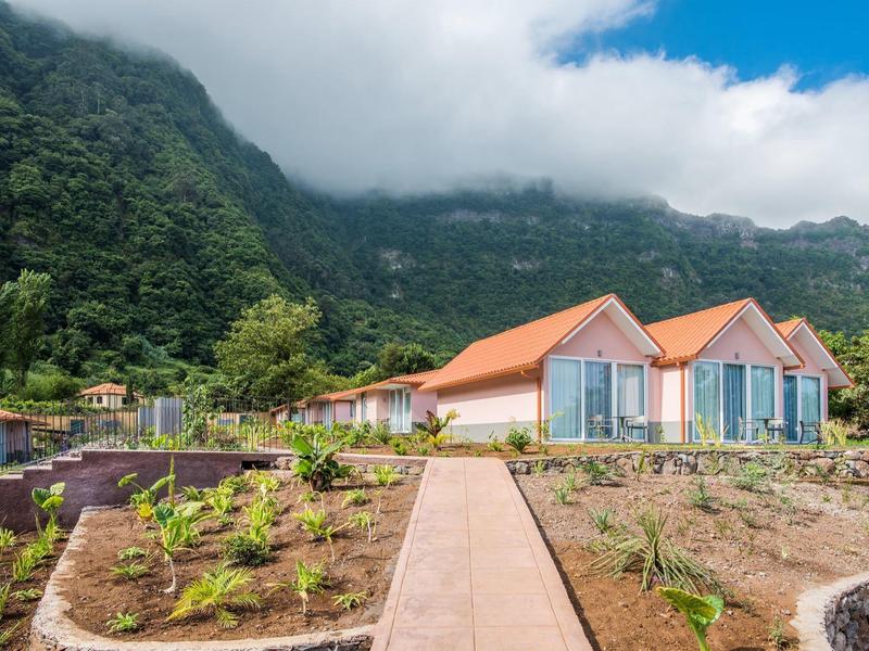 Modern buildings with red roofs in front of forested mountains and cloudy sky.