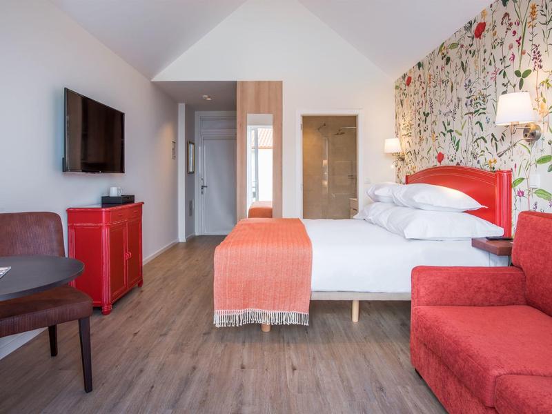 Bright hotel room with double bed, red sofa, wardrobe, TV, and floral wallpaper.