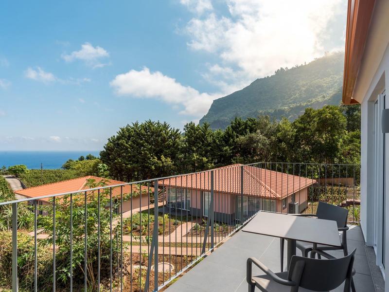 Balcony with chairs and table overlooking sea, mountains, and green vegetation on a sunny day.