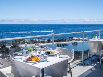 Terrassenrestaurant mit weißen Tischen, Meerblick und sonnigem Himmel im Hintergrund.