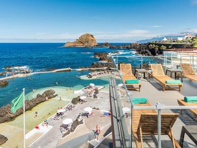 Klares Meer mit Felsen, Sonnenliegen und Menschen am Strand in einer sonnigen Küstenstadt.