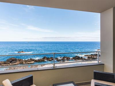Blick auf das blaue Meer mit Felsen am Ufer, gesehen von einem Balkon mit Glasgeländer.
