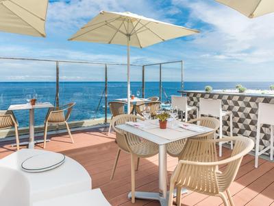 Terrassenrestaurant mit Holzboden, weißen Möbeln, Sonnenschirmen und Blick aufs blaue Meer.