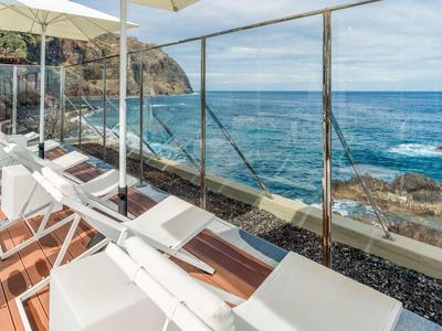 Terrasse mit weißen Liegestühlen und Sonnenschirmen, Blick auf felsige Küste und blaues Meer.