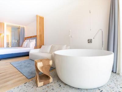 Biała, okrągła wolnostojąca wanna obok sofy i drewnianego stołka w nowoczesnym pokoju hotelowym.