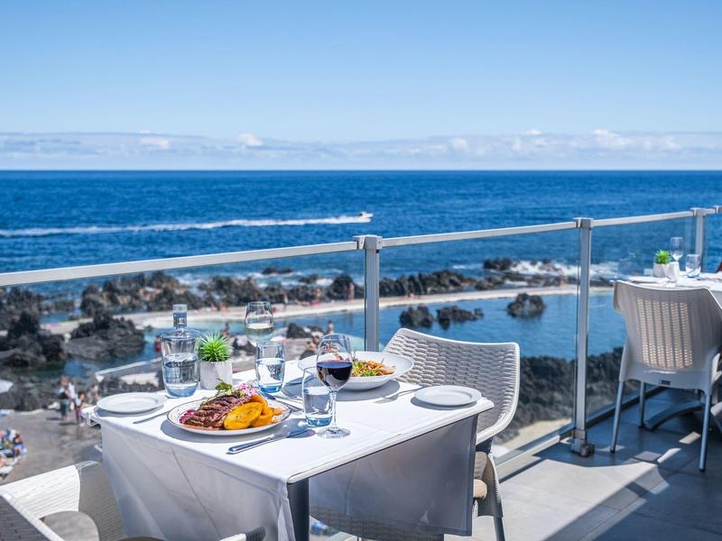 Terrassenrestaurant mit weißen Tischen, Meerblick und sonnigem Himmel im Hintergrund.