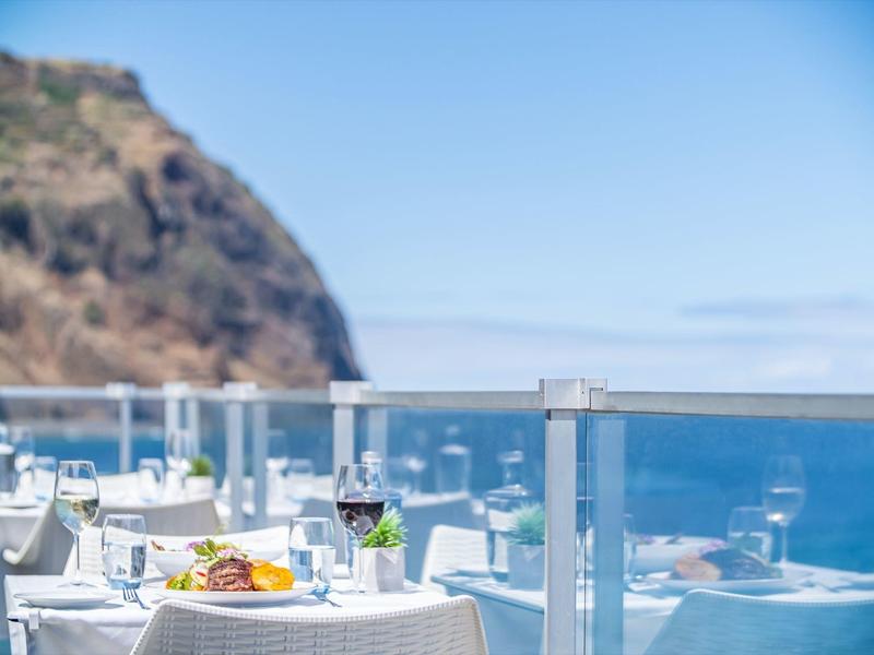 Terrassenrestaurant mit weißen Tischen, Blick auf blaues Meer und Fels im Hintergrund.